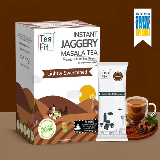 Tea Fit Instant Jaggery Masala Tea premium milk tea premix box shown with a single sachet and a glass of prepared tea on a brown background. The packaging features a stylized rural landscape illustration and text including 'Lightly Sweetened' and 'Premium Milk Tea Premix'.
