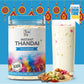 A jar of Tea Fit Instant Thandai Unsweetened powder next to a tall glass filled with the prepared drink, set against a bright blue background with hanging decorative ornaments and a 'As seen on Shark Tank India' badge. Various spices and nuts are scattered at the base.