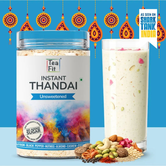 A jar of Tea Fit Instant Thandai Unsweetened powder next to a tall glass filled with the prepared drink, set against a bright blue background with hanging decorative ornaments and a 'As seen on Shark Tank India' badge. Various spices and nuts are scattered at the base.