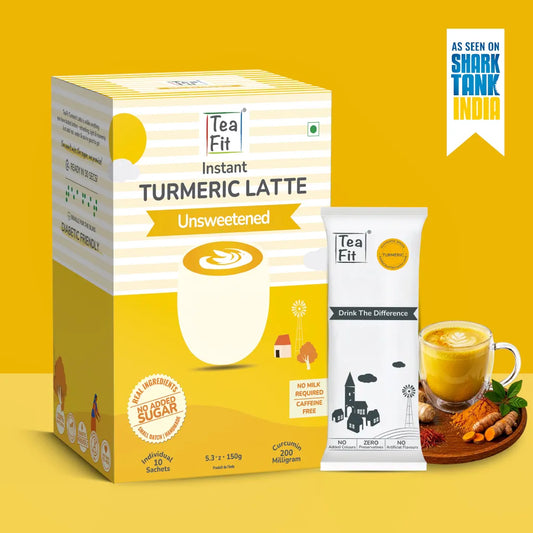 Tea Fit Instant Turmeric Latte Unsweetened product packaging shown with a single sachet, a glass of prepared latte, and turmeric root props on a yellow background. A badge reading 'As seen on Shark Tank India' appears in the top right.
